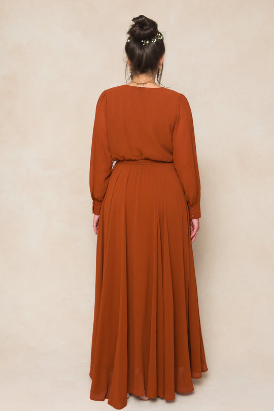 A woman with dark hair styled in a bun and decorated with delicate flowers wears the Ivy City Co Andie Dress in Amber Chiffon—a long-sleeved, floor-length gown—as she stands facing away from the camera against a light beige background.