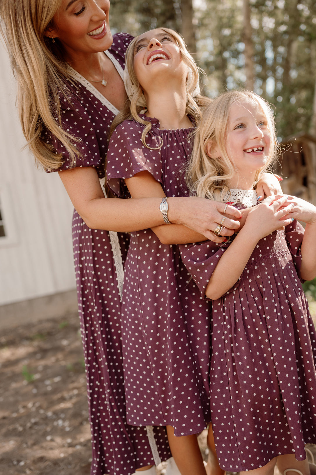 Three smiling, laughing people—a woman and two young girls—stand together outside on a sunny day, all wearing matching Ivy City Co Teen Blakely Dresses in Aubergine Ditsy Dot featuring smocked bodices and flutter sleeves.