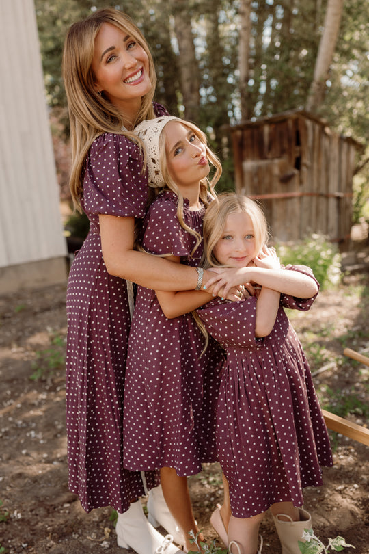 Three smiling females—a woman and two girls—stand outdoors hugging, all dressed in matching Ivy City Co "Teen Blakely Dress in Aubergine Ditsy Dot." The dresses feature flutter sleeves and smocked bodices. Trees and a wooden shed are visible in the background.