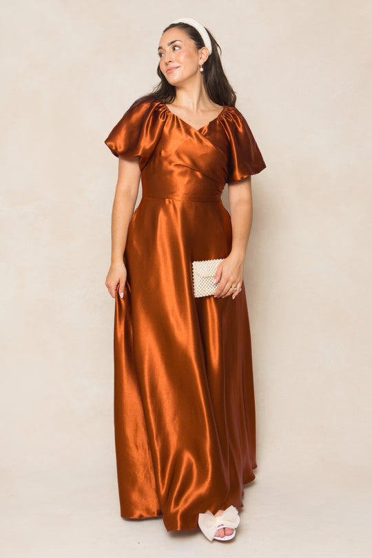 A woman in the Ivy City Co Tessie Dress in Amber Satin, featuring puff sleeves and a shiny finish—ideal for fall bridesmaids—stands against a neutral background, accessorized with a textured clutch, white sandals, and a headband.