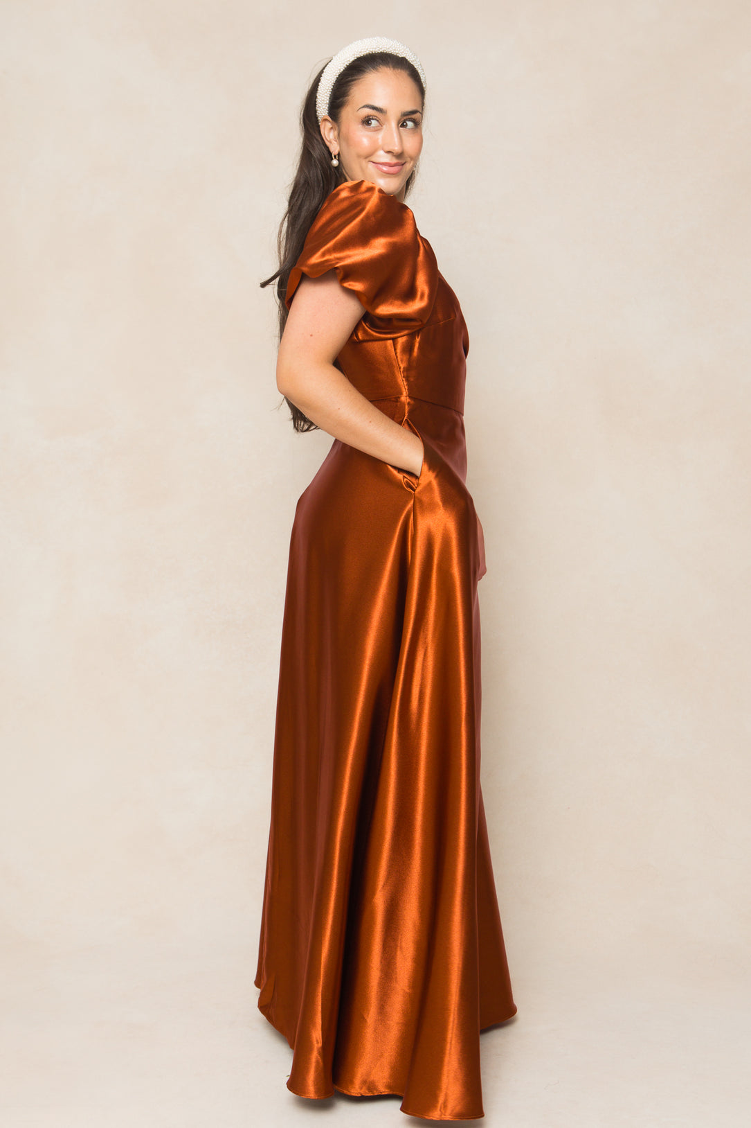 A woman with long dark hair, wearing a white headband and the Tessie Dress in Amber Satin by Ivy City Co, stands sideways with her hands in the dress pockets, smiling softly at the camera.