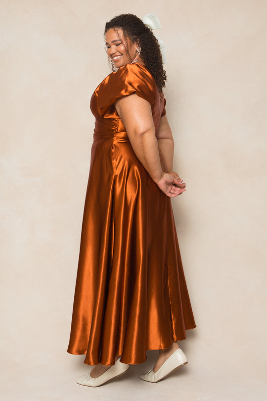 A woman in the Tessie Dress in Amber Satin by Ivy City Co stands sideways, smiling with her hands clasped behind her back. She has curly hair, wears white heels, and poses against a light beige background—perfect for fall bridesmaids.