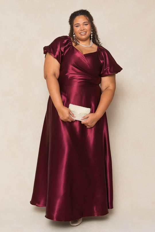 A woman wearing the Ivy City Co Tessie Dress in Wine Satin with short puff sleeves stands holding a beaded clutch and wearing pearl jewelry. Her curly hair and smile make this dress a perfect choice for fall bridesmaids.
