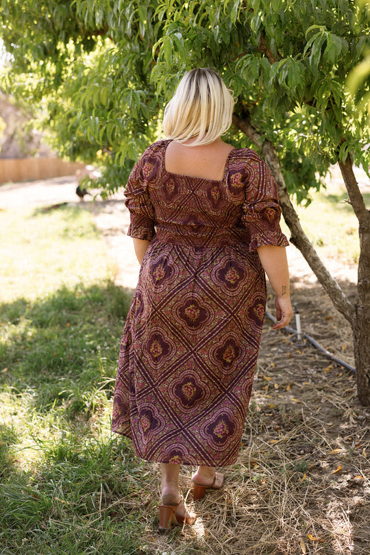 A person with short blonde hair, wearing the Ivy City Co Josephine Dress in Nocturne Diamond Rose and heels, walks away on a grassy path toward a tree on a sunny day.