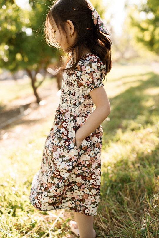 A young girl in the Ivy City Co Mini Sunday Best Dress in Midnight Garden, featuring puff sleeves and a smocked bodice, stands sideways in a sunlit grassy area with trees behind her. She has her hands in her pockets and wears a matching hair bow.