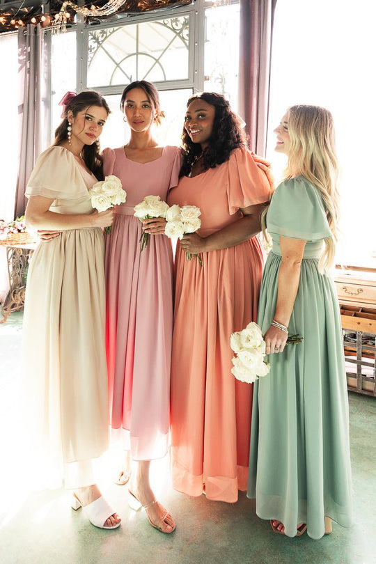 Four women in pastel-colored dresses, including the Abbie Dress in Blush by Ivy City Co—a modest bridesmaid dress with flutter sleeves—hold white bouquets as they stand together indoors by a window with soft natural light. They are smiling and appear to be bridesmaids at a wedding.