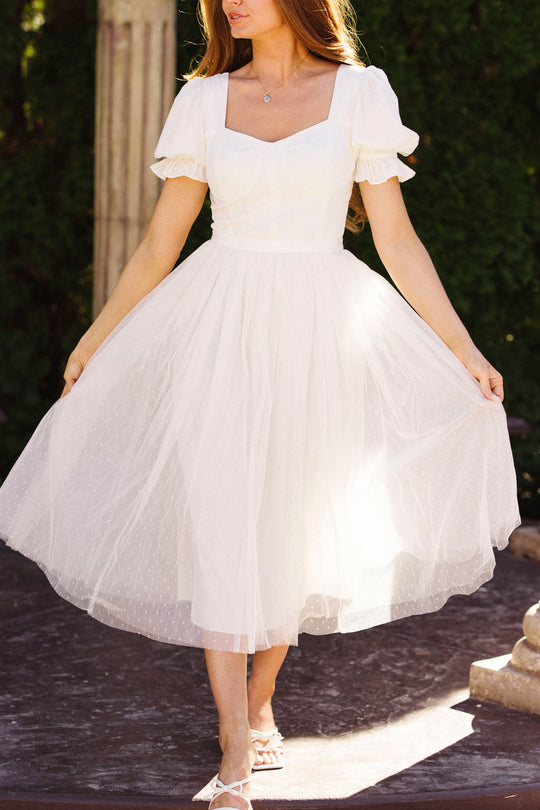A woman wears the Ivy City Co Ballerina Dress in White, featuring a fitted bodice and flared tulle skirt. The light, short-sleeved style adds charm as she stands outdoors, holding the sides of her dress with her face partly out of frame.