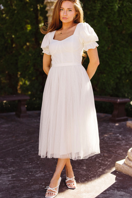 A woman stands outdoors in the Ivy City Co Ballerina Dress in White, featuring a fitted bodice and midi tulle skirt. She wears white strappy sandals, with greenery and stone benches in the background.
