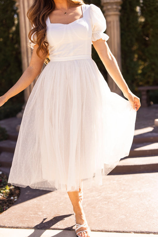 A woman stands outdoors in the Ivy City Co Ballerina Dress in White, featuring short sleeves and a square neckline. Sunlight illuminates the delicate tulle fabric as she holds her skirt, her long wavy hair catching the light.