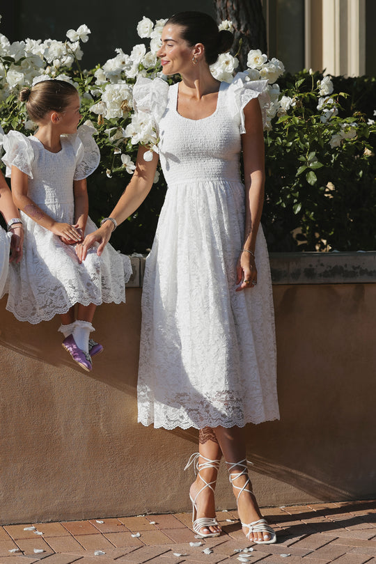 A woman and a young girl wear matching Ivy City Co Hattie Dresses in White Lace, standing outdoors by white flowers. The elegant, romantic dresses are perfect for a summery day.