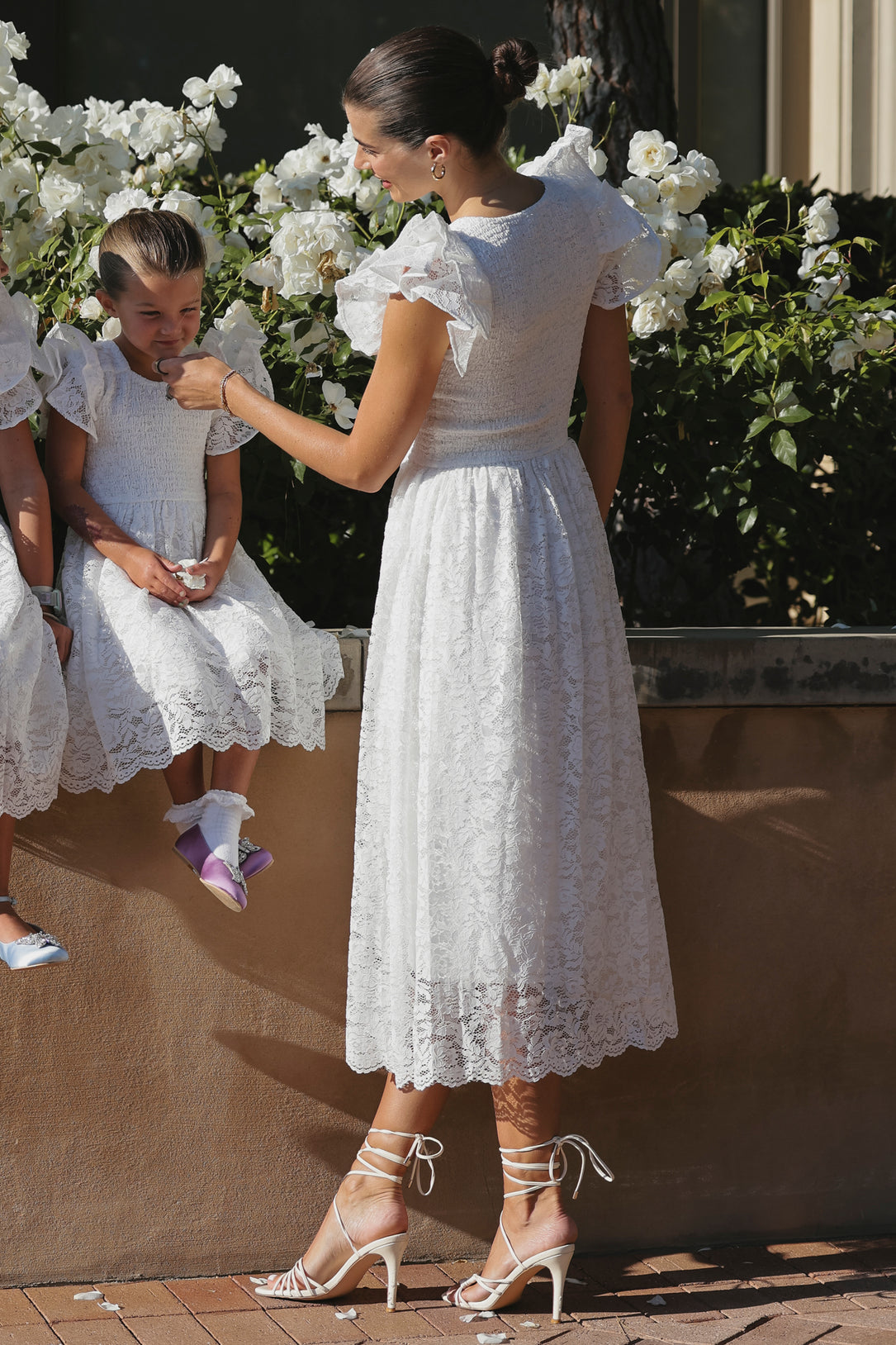 A woman in the Ivy City Co Hattie Dress in White Lace and strappy heels helps a young girl, also wearing the Hattie Dress, adjust her hair by a low wall with blooming white flowers.