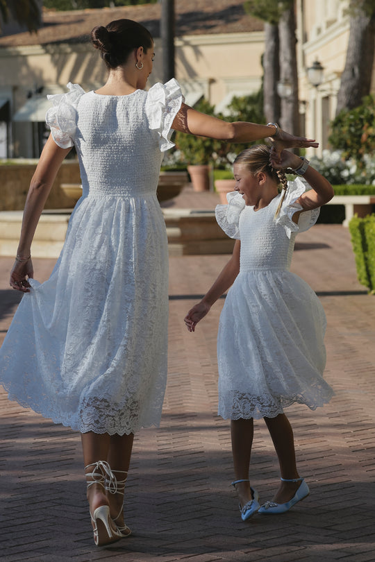 A woman and young girl dance outdoors in matching Ivy City Co Hattie Dresses in White Lace, holding hands as they twirl along a path with greenery, their romantic dresses fluttering in the sunny breeze.