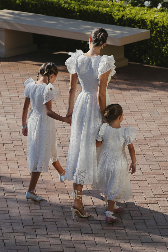 A woman and two young girls wear the Ivy City Co Hattie Dress in White Lace with sandals, walking hand in hand on a sunlit brick path, greenery and benches behind them.