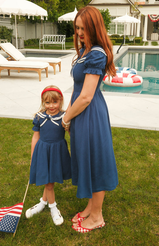 A woman and a young girl wear matching Ivy City Co Alice Dresses in Sailor Navy, standing by the grass near a pool. The girl holds an American flag as they enjoy a sunny, maternity-friendly outdoor scene.
