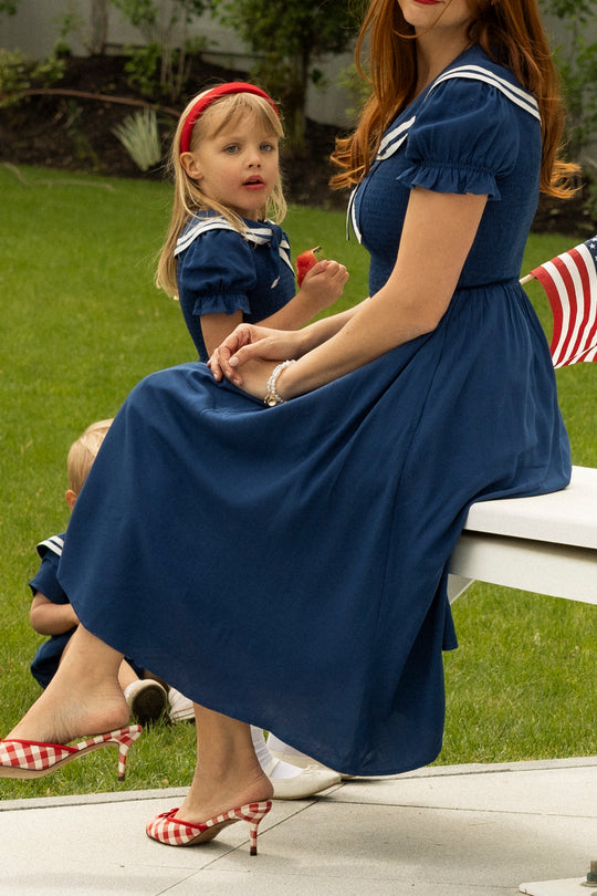 A woman in an Ivy City Co Alice Dress in Sailor Navy sits on grass with two young girls in matching dresses; one wears a red headband, and the woman pairs her outfit with red gingham heels.
