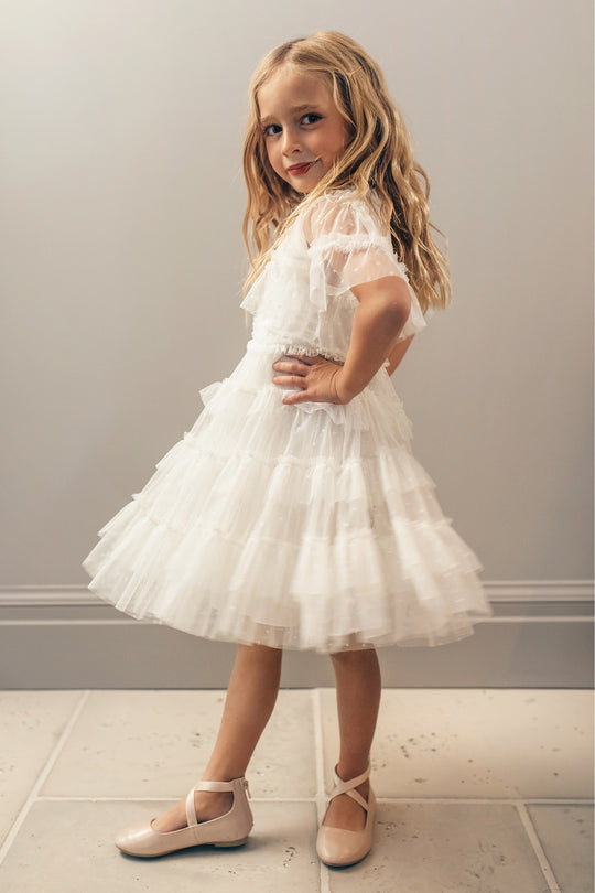 A young girl with long blonde hair wears the Ivy City Co Mini Whimsical Dress in White and light pink ballet flats, standing sideways with one hand on her hip and smiling confidently in front of a plain light-colored wall.
