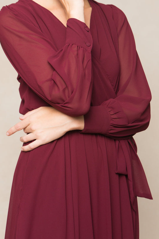 A person models the Andie Dress in Wine Chiffon by Ivy City Co, featuring elegant long sleeves and a flowing maxi silhouette. With one arm crossed and the other hand touching their chin, the photo highlights the sophisticated bridesmaids dress and graceful hand pose, cropped above the neck for emphasis.
