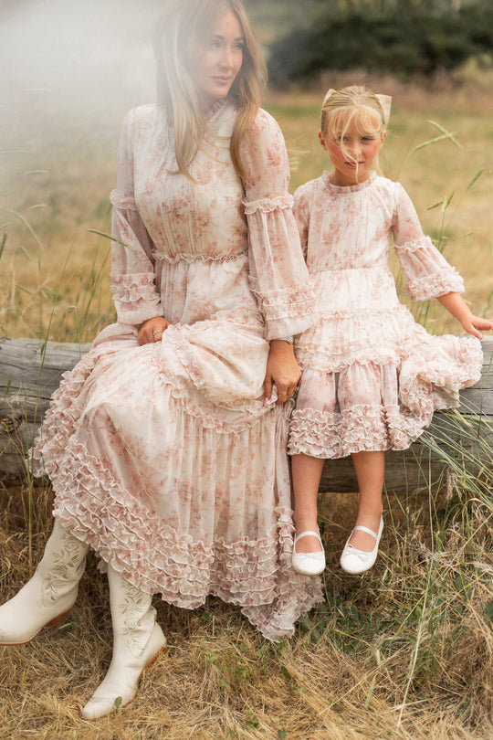 A woman and a young girl sit on a log in a grassy field, both wearing matching Ivy City Co Geneva Dresses in Banana Creme Aquarelle Fleur. The woman styles hers with white boots; the girl adds white shoes and sunglasses to her ruffled look.