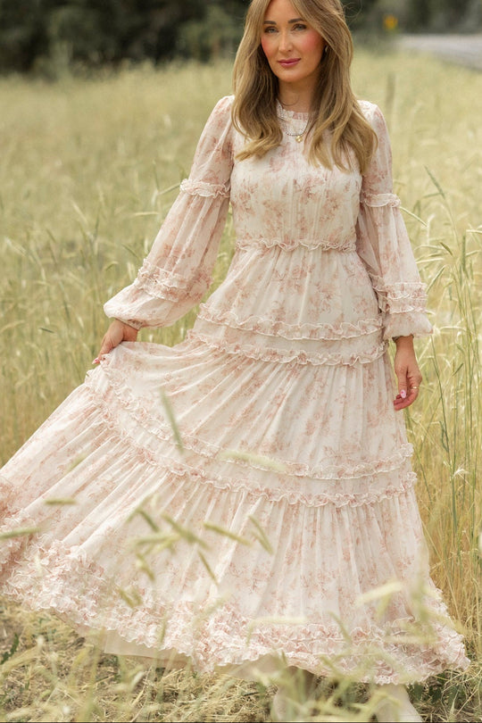 A woman stands in a grassy field wearing the Geneva Dress in Banana Creme Aquarelle Fleur by Ivy City Co, gently holding one side of her flowing floral dress with ruffle details as she looks at the camera, surrounded by tall grass.