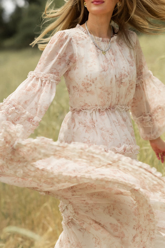 A woman twirls outdoors in the Ivy City Co Geneva Dress in Banana Creme Aquarelle Fleur, her hair and dress billowing as she moves amid tall grass and soft greenery.