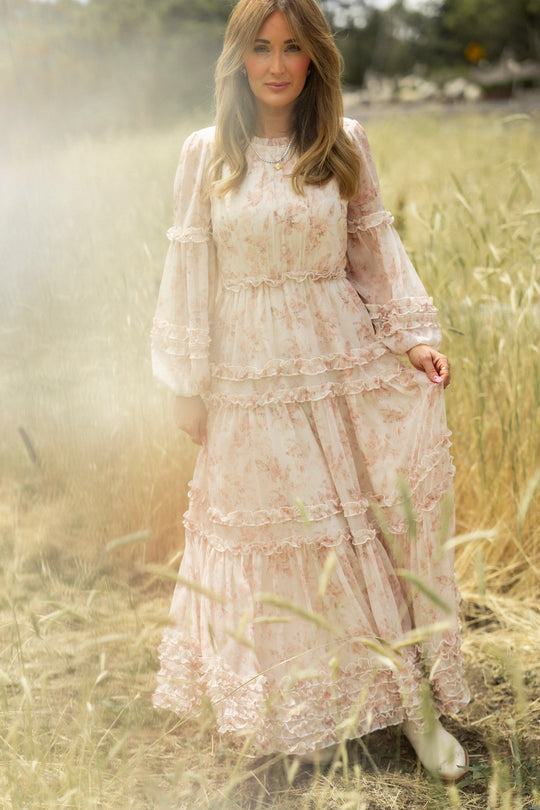 A woman with light brown hair stands in a dreamy, sunlit field, holding the Geneva Dress in Banana Creme Aquarelle Fleur by Ivy City Co, its watercolor floral print and ruffles creating an ethereal look as she gazes at the camera.