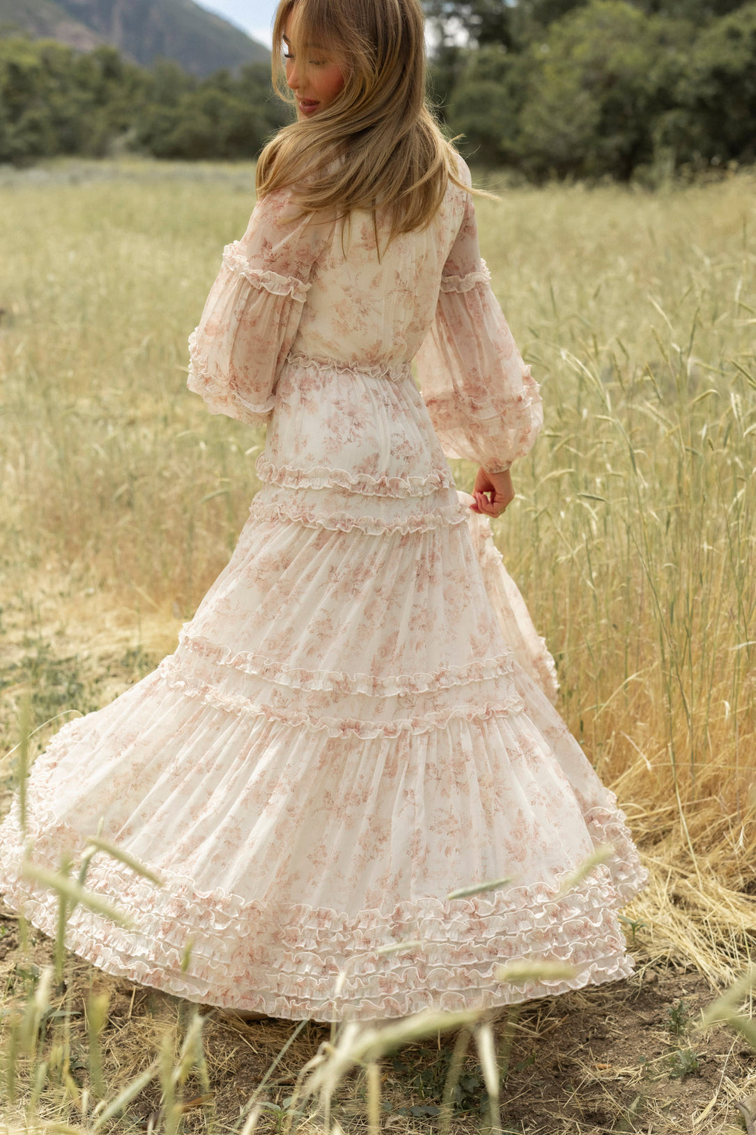A woman with long light brown hair wears the Geneva Dress in Banana Creme Aquarelle Fleur by Ivy City Co, standing in a grassy field and looking over her shoulder as the elegant dress contrasts with trees and mountains behind her.