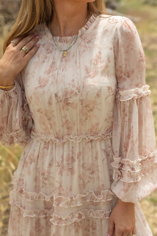 A woman with wavy light brown hair stands outdoors, wearing the Ivy City Co Geneva Dress in Banana Creme Aquarelle Fleur, a long-sleeved, ruffled watercolor floral design, accessorized with rings, bracelets, and a layered necklace.