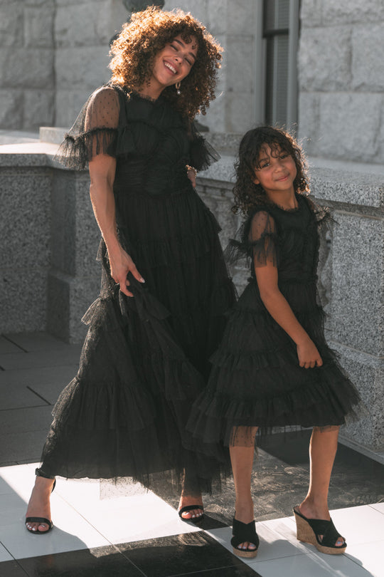 A woman and a young girl smile outdoors, both wearing matching Mini Whimsical Dresses in Black from Ivy City Co. Their layered tulle skirts and open-toed heels catch the sunlight as they stand close together by a stone wall, radiating joy.