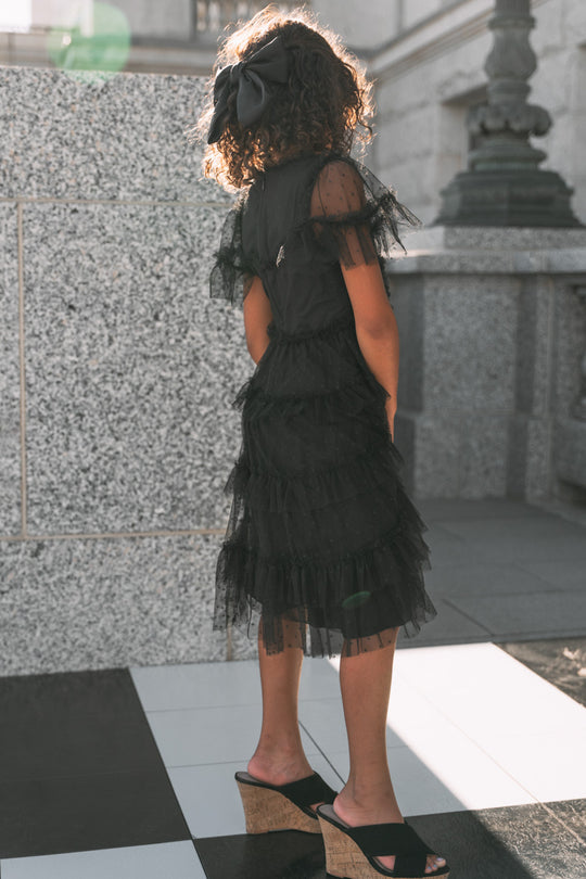 A child wearing the Ivy City Co Mini Whimsical Dress in Black, featuring a frilly layered tulle skirt, pairs the outfit with a large hair bow and black wedge sandals while standing outdoors on a tiled floor near a sunlit stone wall, facing away.