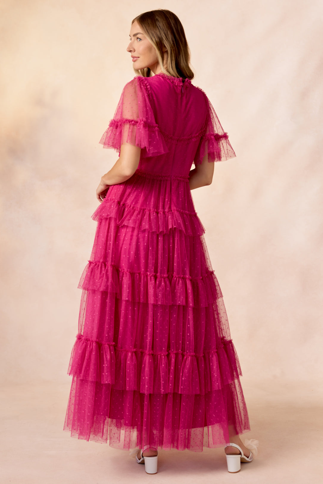 A woman with shoulder-length hair and white heeled sandals faces away, wearing the Ivy City Co Whimsical Dress in Fuchsia—featuring tiered tulle, ruffled sleeves, and polka dot details—set against a soft, light backdrop.