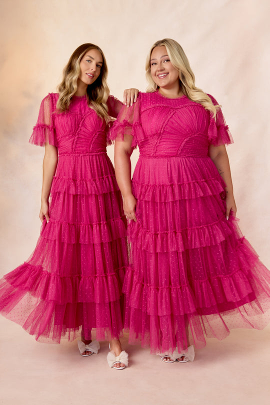 Two women smile side by side, wearing matching Whimsical Dress in Fuchsia by Ivy City Co, featuring ruffled tiered tulle skirts and sheer short sleeves, paired with white shoes against a soft, light background.
