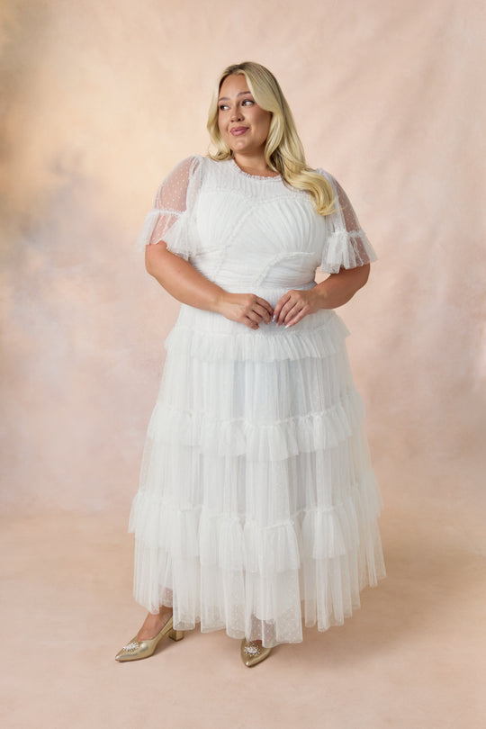 A woman with long blonde hair wears the Ivy City Co Whimsical Dress in White, featuring a tulle skirt and flutter sleeves, paired with gold flats, standing against a beige and pink backdrop.
