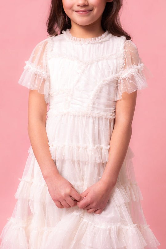 A young girl wearing the Ivy City Co Mini Whimsical Dress in White, featuring sheer ruffled sleeves and a layered tulle skirt, stands against a pink background, smiling softly with her hands clasped in front of her.