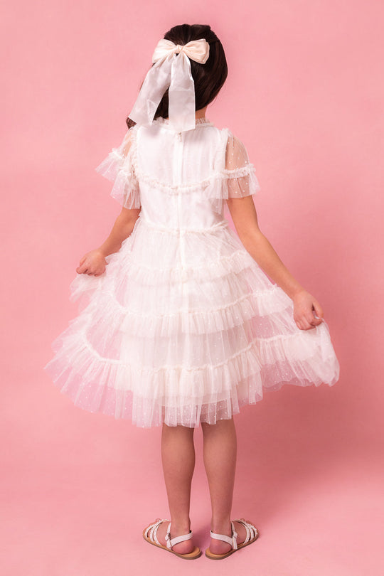 A girl wearing the Ivy City Co Mini Whimsical Dress in White—with its ruffled details and layered tulle skirt—stands against a pink background, facing away. She has a large white bow in her hair, holds the sides of her dress, and completes the look with sandals.