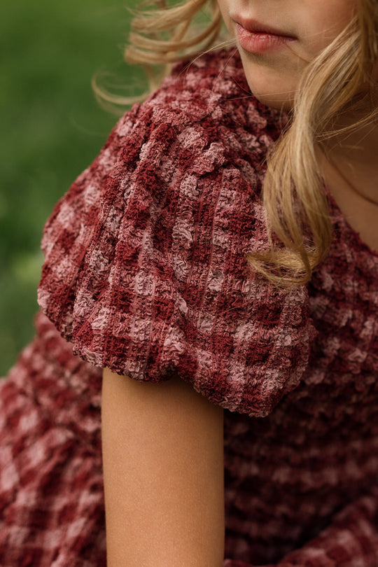 A young girl with blonde curly hair sits outdoors on green grass, wearing the Ivy City Co Mini Tori Dress in Cranberry Popcorn Plaid; the image highlights her arm, a partial glimpse of her face, and showcases the dress’s charming puff sleeves and smocked bodice.