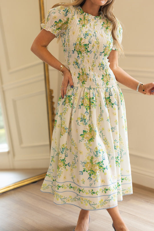 A woman wears the Jennifer Dress in Garden Tea Party Floral by Ivy City Co—a white, short-sleeve dress with a smocked, gathered waist and lightweight fabric adorned with yellow and green floral patterns. She stands indoors with only her body visible, not her face.