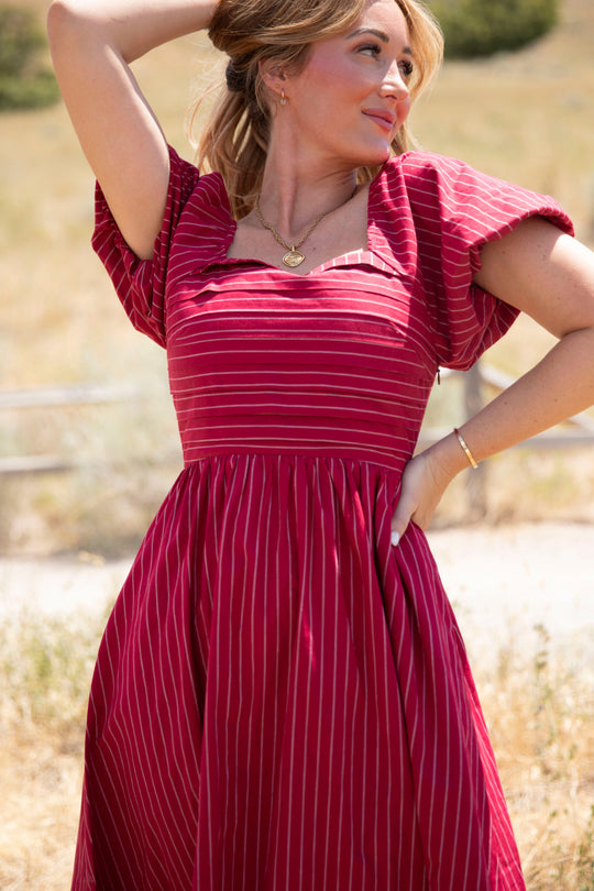 A woman wearing the Ivy City Co Julie Dress in Cranberry Pinstripe—with puff sleeves and a smocked bodice—stands outdoors in a sunlit grassy area, gazing to the side with one hand behind her head and the other on her hip, against a blurred backdrop of trees.