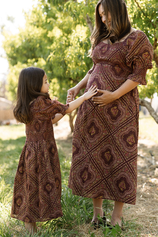 A pregnant woman and a young girl, both wearing matching Ivy City Co Josephine Dresses in Nocturne Diamond Rose with smocked bodices, smile while touching the woman’s belly outdoors among green trees.