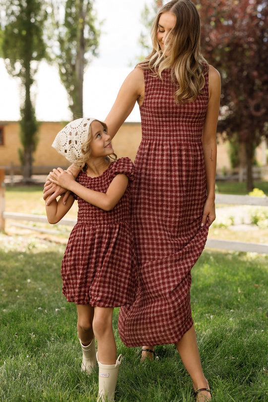 A woman and a young girl, both dressed in the Ivy City Co Mini Tori Dress in Cranberry Popcorn Plaid, smile at each other while holding hands and standing on green grass outdoors. The girl’s outfit is completed with a white knit hat and boots.