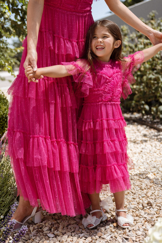 A smiling young girl wears the Ivy City Co Mini Whimsical Dress in Fuchsia, featuring a layered tulle skirt and flutter sleeves, as she stands on rocks holding hands with an adult in a matching pink dress. Green plants provide a lush background.