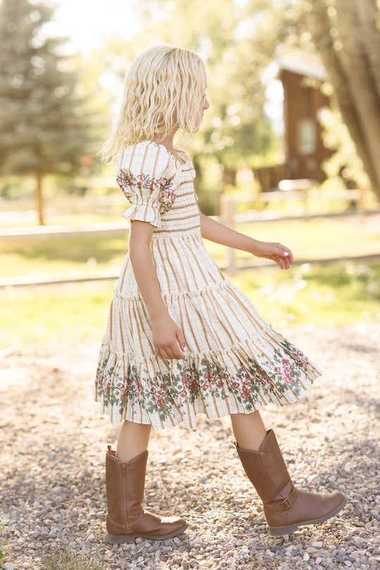 A young child with blonde hair walks along a gravel path outside, sunlight streaming through the trees, wearing the Ivy City Co Mini Madeline Dress in Embroidered Cider Grid, which features a smocked bodice and charming puff sleeves.