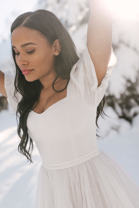 A woman in the Ivy City Co Ballerina Dress in White stands outdoors in a snowy landscape, eyes closed and arms raised as sunlight creates a soft, serene atmosphere.
