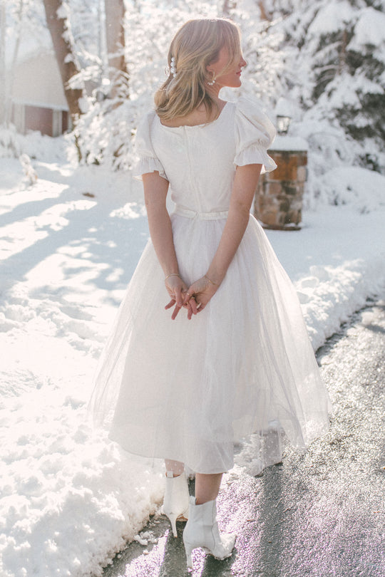 A woman wearing the Ivy City Co Ballerina Dress in White and white boots stands on a snowy path, hands clasped behind her, facing sunlit snow-covered trees.