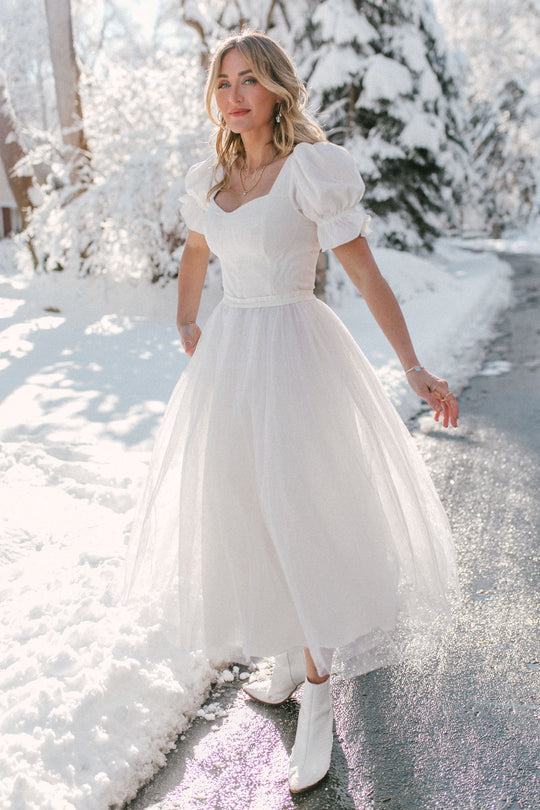 A woman wears the Ivy City Co Ballerina Dress in White with boots, standing on a snowy path surrounded by sunlit, snow-covered trees.