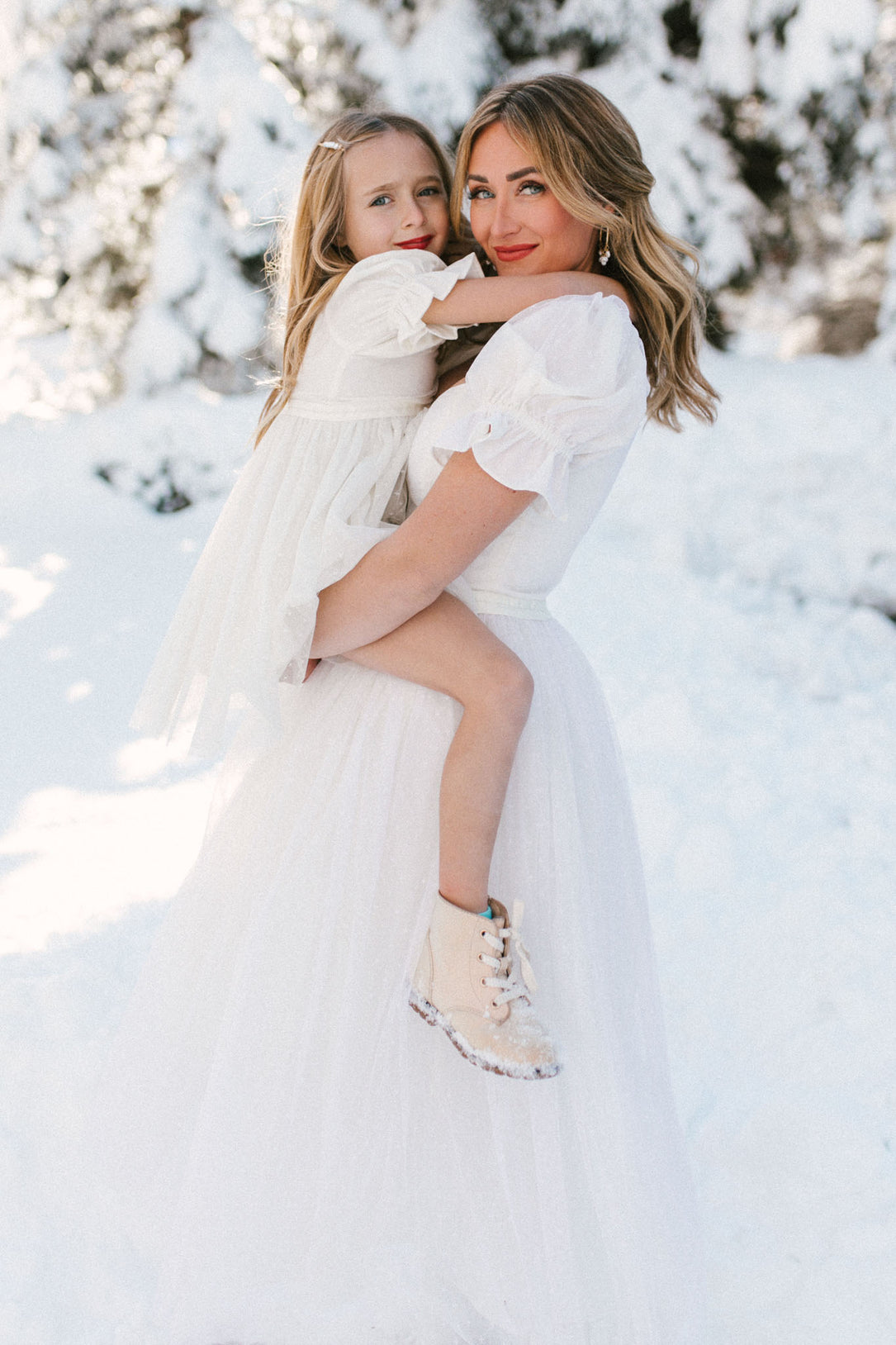 A woman and a young girl, both smiling, wear matching Ivy City Co Ballerina Dresses in White while standing outdoors among snow-covered trees.