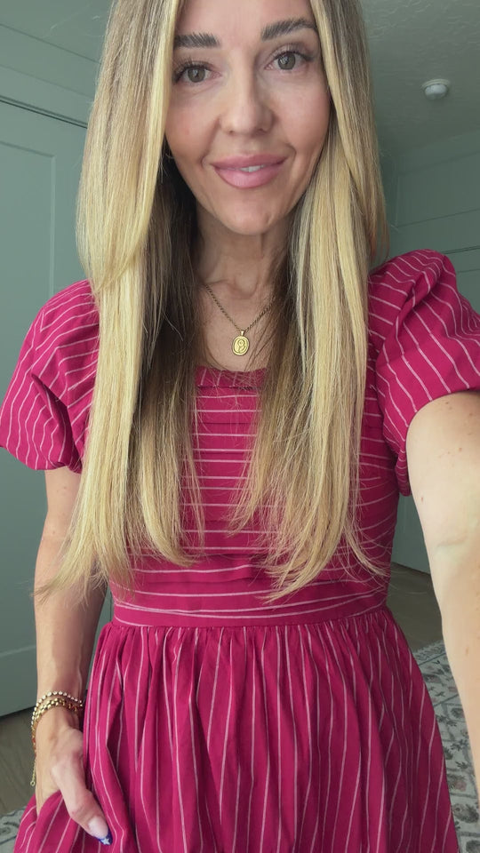 A smiling woman with long blonde hair wears the Ivy City Co Julie Dress in Cranberry Pinstripe, featuring a smocked bodice and puff sleeves, standing indoors.