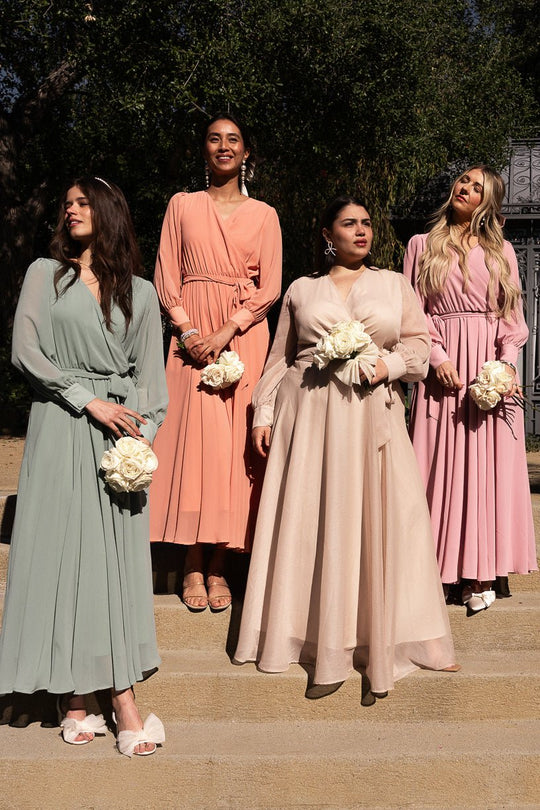 Four women in pastel-colored dresses, including the Ivy City Co Andie Dress in Blush, stand on outdoor steps, each holding a bouquet of white flowers. Sunlight highlights their outfits, while trees and a wrought iron gate are visible in the background.