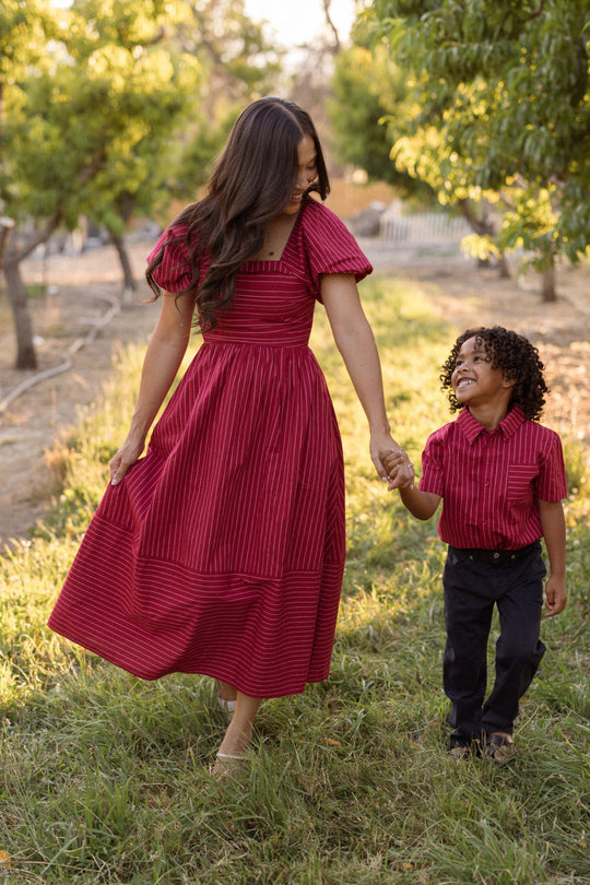 Wearing the Ivy City Co Julie Dress in Cranberry Pinstripe, a woman with puff sleeves and a smocked bodice holds hands and walks with a young boy in a red shirt and dark pants through a grassy area with trees, both smiling at each other.