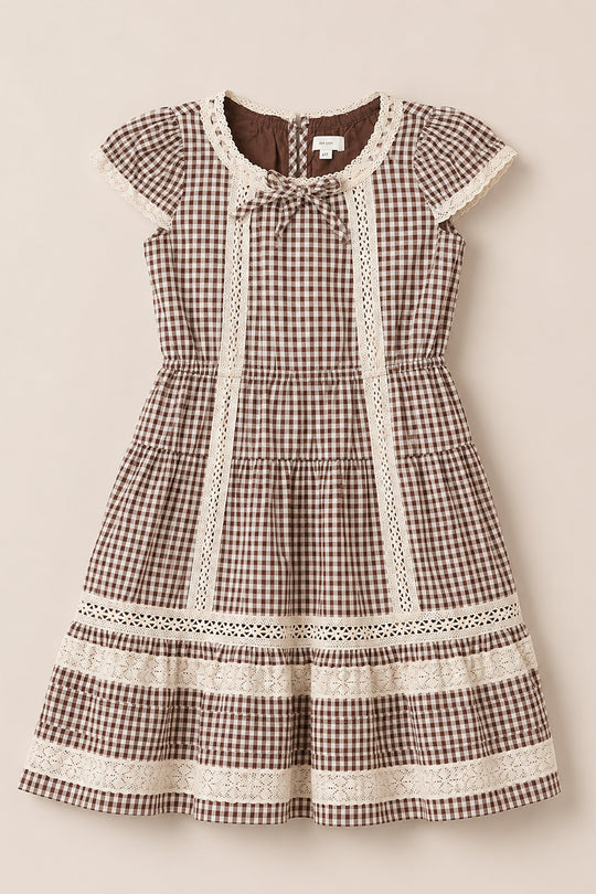 The Mini Palermo Dress in Hickory Gingham by Ivy City Co is a brown and white gingham girls dress featuring short sleeves, lace trim details, a round neckline adorned with a small bow, and a flared skirt, crafted from soft cotton-blend fabric—shown laid flat on a neutral background.