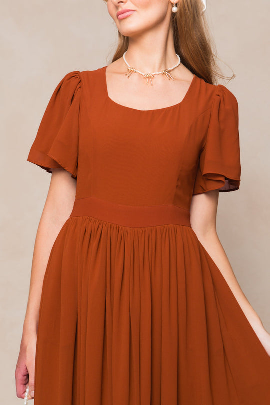 A woman poses against a neutral background wearing the Ivy City Co Abbie Dress in Amber Chiffon—a rust-colored, short-sleeve dress with a square neckline and gathered skirt reminiscent of a classic chiffon bridesmaid dress—paired with a pearl necklace featuring a bow detail.